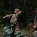 Two soldiers in US Army OCP (Operational Camouflage Pattern) uniforms are engaged in what appears to be a field training exercise in a wooded area. The soldier on the left, wearing a helmet and tactical vest, is in a throwing motion, likely practicing grenade throwing. A second soldier stands nearby observing, possibly acting as a safety observer or instructor.