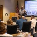A professional presentation is taking place in a conference room where a man in a gray suit is speaking at a podium about 'OC-ALC Workforce Development.' The projected slide details collaboration with State Career Tech Centers, Secondary Education Sites, and Oklahoma State University development programs. An audience of approximately 10-15 attendees is seated at conference tables facing the presenter.