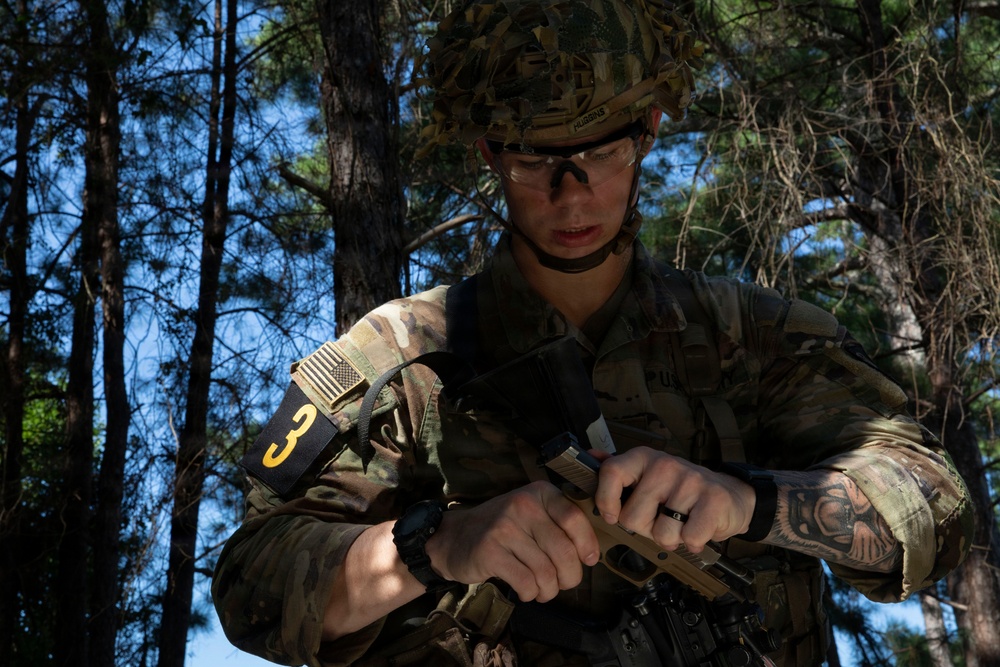 A U.S. Army soldier in full multicam combat gear is handling a pistol, likely performing a magazine change or weapons check in a wooded training environment. The soldier wears a combat helmet, protective eyewear, body armor, and displays a number '3' identifier on their arm along with a reversed U.S. flag patch. The setting appears to be a field training exercise based on the forested background and competition-style numbering.