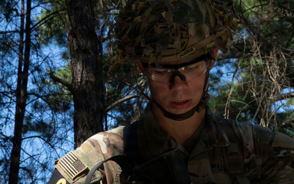 A U.S. Army soldier in full multicam combat gear is handling a pistol, likely performing a magazine change or weapons check in a wooded training environment. The soldier wears a combat helmet, protective eyewear, body armor, and displays a number '3' identifier on their arm along with a reversed U.S. flag patch. The setting appears to be a field training exercise based on the forested background and competition-style numbering.