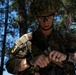 A U.S. Army soldier in full multicam combat gear is handling a pistol, likely performing a magazine change or weapons check in a wooded training environment. The soldier wears a combat helmet, protective eyewear, body armor, and displays a number '3' identifier on their arm along with a reversed U.S. flag patch. The setting appears to be a field training exercise based on the forested background and competition-style numbering.