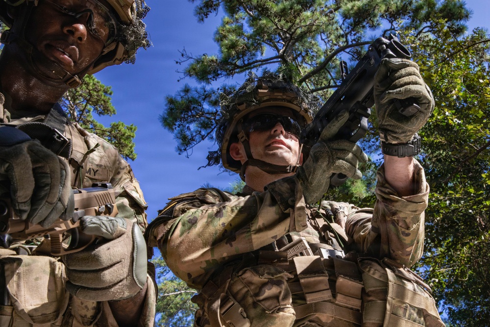 Two U.S. Army soldiers in full combat gear are photographed from a low angle against a blue sky with pine trees. The soldier in the foreground is raising a pistol upward while wearing tactical gloves, sunglasses, and a camouflage helmet. Both soldiers wear OCP (Operational Camouflage Pattern) uniforms with visible 'ARMY' tape and plate carrier vests.