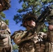 Two U.S. Army soldiers in full combat gear are photographed from a low angle against a blue sky with pine trees. The soldier in the foreground is raising a pistol upward while wearing tactical gloves, sunglasses, and a camouflage helmet. Both soldiers wear OCP (Operational Camouflage Pattern) uniforms with visible 'ARMY' tape and plate carrier vests.