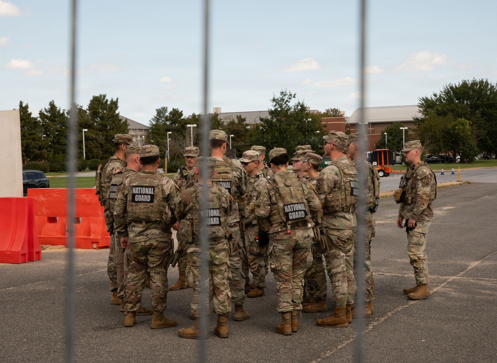 A group of approximately 10-12 National Guard soldiers in camouflage uniforms are gathered in a parking lot, appearing to receive a briefing or conduct a formation huddle. The image is photographed through vertical metal fence or barrier bars in the foreground. Orange traffic barriers and a brick building with trees are visible in the background.