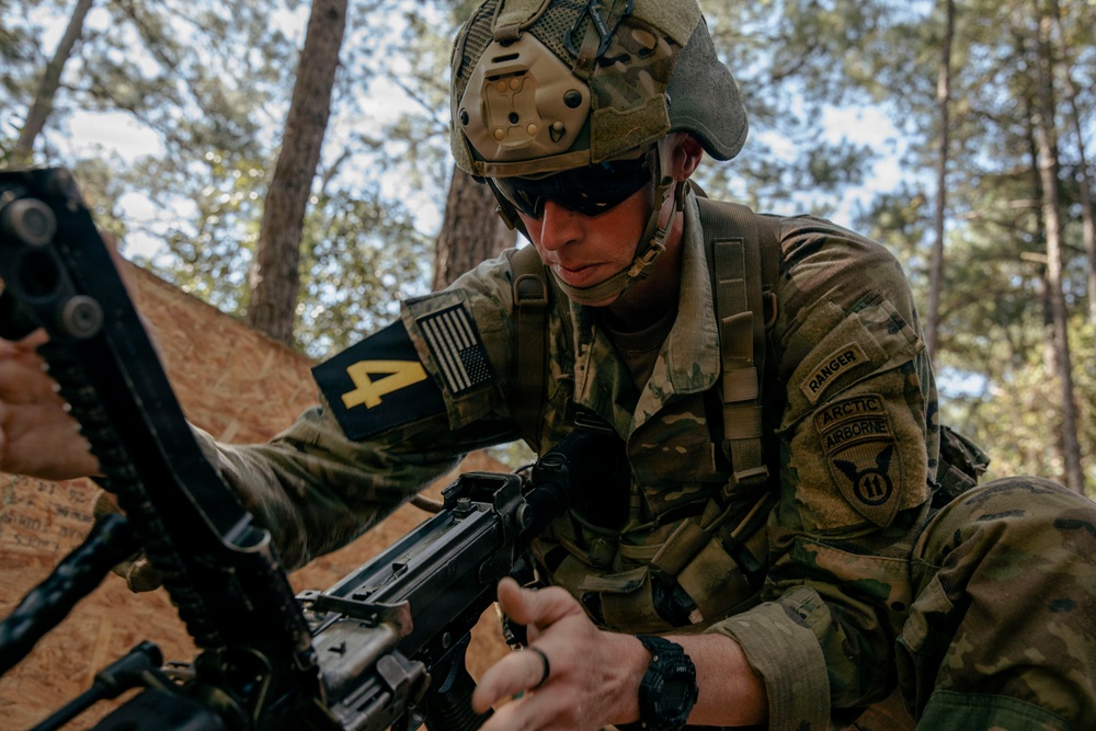 A U.S. Army soldier in full combat gear is handling a weapon in a wooded training environment. The soldier displays multiple unit patches including Ranger, Arctic, Airborne, and 11th Airborne Division insignia on their OCP (Operational Camouflage Pattern) uniform. The image appears to capture a field training exercise or weapons handling drill.