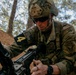 A U.S. Army soldier in full combat gear is handling a weapon in a wooded training environment. The soldier displays multiple unit patches including Ranger, Arctic, Airborne, and 11th Airborne Division insignia on their OCP (Operational Camouflage Pattern) uniform. The image appears to capture a field training exercise or weapons handling drill.