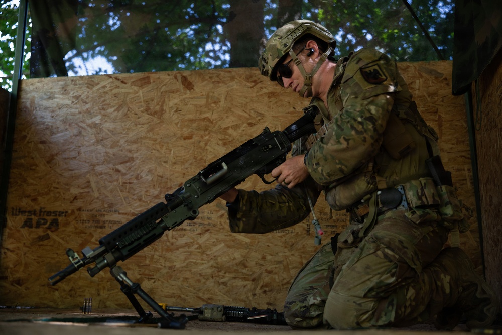 A soldier in full multicam combat gear is kneeling on the ground while handling a light machine gun with a bipod deployed. The setting appears to be a training environment constructed with plywood walls. The soldier is equipped with a tactical helmet, ballistic sunglasses, body armor, and communications equipment.