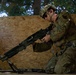 A soldier in full multicam combat gear is kneeling on the ground while handling a light machine gun with a bipod deployed. The setting appears to be a training environment constructed with plywood walls. The soldier is equipped with a tactical helmet, ballistic sunglasses, body armor, and communications equipment.