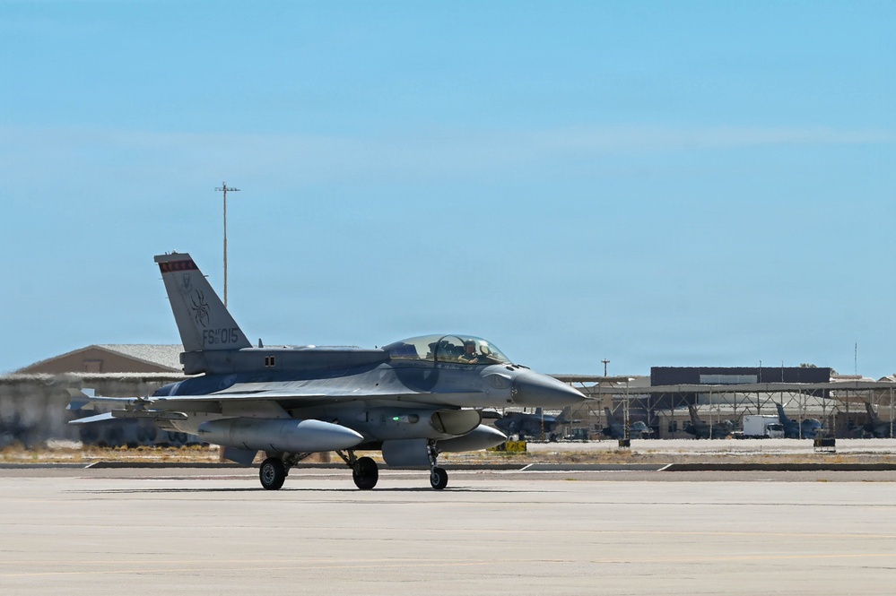 A single-seat F-16 Fighting Falcon fighter jet is taxiing on a concrete runway apron at a military air base. The aircraft bears tail code 'FS 015' and displays a unit insignia on the vertical stabilizer. Military hangars and support structures are visible in the background under clear blue skies.
