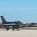 A single-seat F-16 Fighting Falcon fighter jet is taxiing on a concrete runway apron at a military air base. The aircraft bears tail code 'FS 015' and displays a unit insignia on the vertical stabilizer. Military hangars and support structures are visible in the background under clear blue skies.