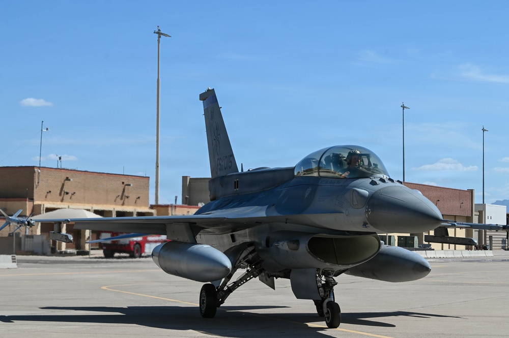 A single-seat F-16 Fighting Falcon jet fighter is taxiing on a military airbase flight line. The aircraft is painted in overall gray livery and has a pilot visible in the cockpit. The setting appears to be a U.S. Air Force installation, likely in a desert environment based on the terrain and architecture visible in the background.