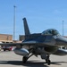 A single-seat F-16 Fighting Falcon jet fighter is taxiing on a military airbase flight line. The aircraft is painted in overall gray livery and has a pilot visible in the cockpit. The setting appears to be a U.S. Air Force installation, likely in a desert environment based on the terrain and architecture visible in the background.
