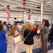 A group of approximately 20 civilians and military personnel are gathered inside what appears to be a military facility or base, likely during an official tour or briefing. Two American flags are prominently displayed hanging from the ceiling of the large industrial-style building. The group includes a mix of business-attired civilians and uniformed military members, suggesting a community outreach, employer support, or official visit event.