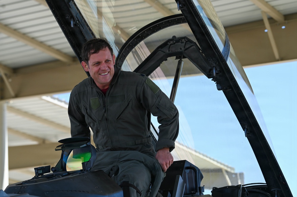A male military pilot in an olive/sage green flight suit is seated in the open cockpit of a military jet aircraft, smiling broadly. The aircraft appears to be parked under a covered hangar or shelter structure. The image captures a candid, positive moment likely associated with a flight or post-flight activity.