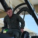 A male military pilot in an olive/sage green flight suit is seated in the open cockpit of a military jet aircraft, smiling broadly. The aircraft appears to be parked under a covered hangar or shelter structure. The image captures a candid, positive moment likely associated with a flight or post-flight activity.