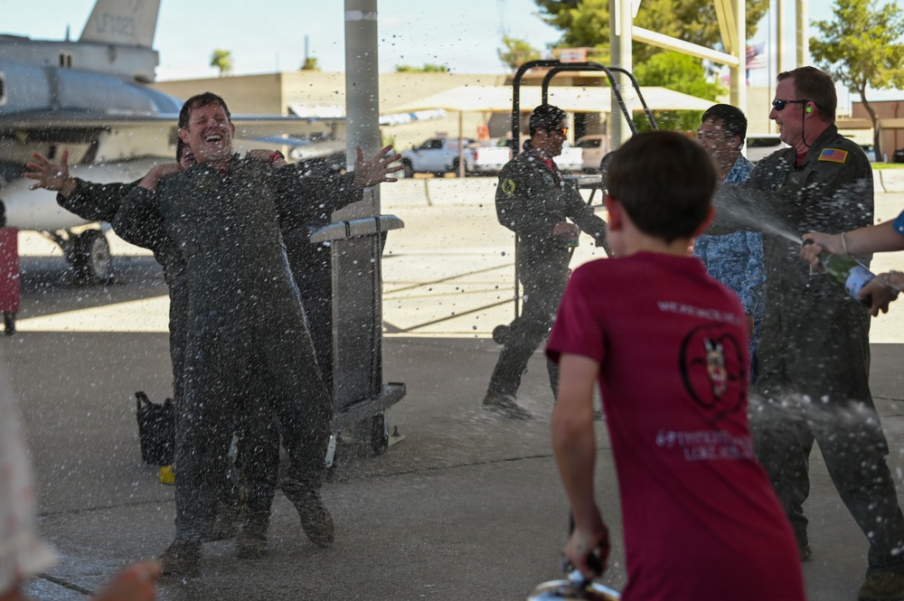A military pilot in a flight suit is being doused with water in what appears to be a traditional 'fini flight' or solo celebration ceremony at a military airbase. A young boy in a red t-shirt is spraying water at the laughing pilot, while other personnel in flight suits observe in the background. An F-16 fighter jet is visible in the background, confirming the military aviation setting.