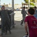 A military pilot in a flight suit is being doused with water in what appears to be a traditional 'fini flight' or solo celebration ceremony at a military airbase. A young boy in a red t-shirt is spraying water at the laughing pilot, while other personnel in flight suits observe in the background. An F-16 fighter jet is visible in the background, confirming the military aviation setting.