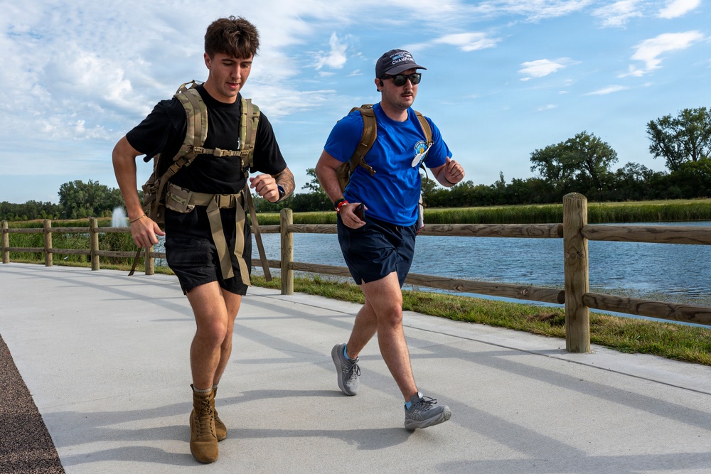 Two men are running or rucking along a paved path near a body of water, both wearing tactical-style backpacks. The setting appears to be a park or recreational trail on a partly cloudy day. Their attire and loaded packs suggest a fitness challenge, military-style ruck march, or charity event.
