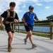 Two men are running or rucking along a paved path near a body of water, both wearing tactical-style backpacks. The setting appears to be a park or recreational trail on a partly cloudy day. Their attire and loaded packs suggest a fitness challenge, military-style ruck march, or charity event.