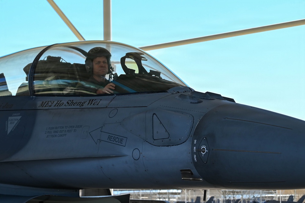 A pilot is seated in the cockpit of a military fighter jet, identified as an F-16 Fighting Falcon based on its distinctive design features. The aircraft bears the name 'ME2 Ho Sheng Wei' stenciled below the canopy, suggesting a Taiwanese or allied air force context. The pilot is wearing a flight helmet with communications equipment and appears to be conducting pre-flight or ground operations.