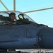 A pilot is seated in the cockpit of a military fighter jet, identified as an F-16 Fighting Falcon based on its distinctive design features. The aircraft bears the name 'ME2 Ho Sheng Wei' stenciled below the canopy, suggesting a Taiwanese or allied air force context. The pilot is wearing a flight helmet with communications equipment and appears to be conducting pre-flight or ground operations.