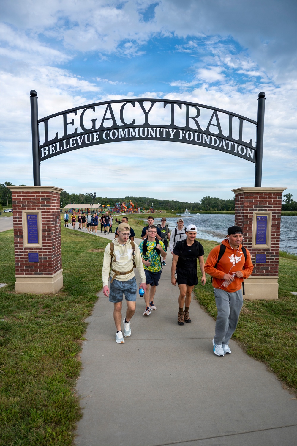 A group of young people walk toward the camera under a large decorative arch reading 'Legacy Trail – Bellevue Community Foundation.' The setting is a paved recreational trail adjacent to a lake, with a playground and fountain visible in the background. The scene appears to be a group walk or organized outdoor activity on a partly cloudy summer day.