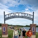 A group of young people walk toward the camera under a large decorative arch reading 'Legacy Trail – Bellevue Community Foundation.' The setting is a paved recreational trail adjacent to a lake, with a playground and fountain visible in the background. The scene appears to be a group walk or organized outdoor activity on a partly cloudy summer day.