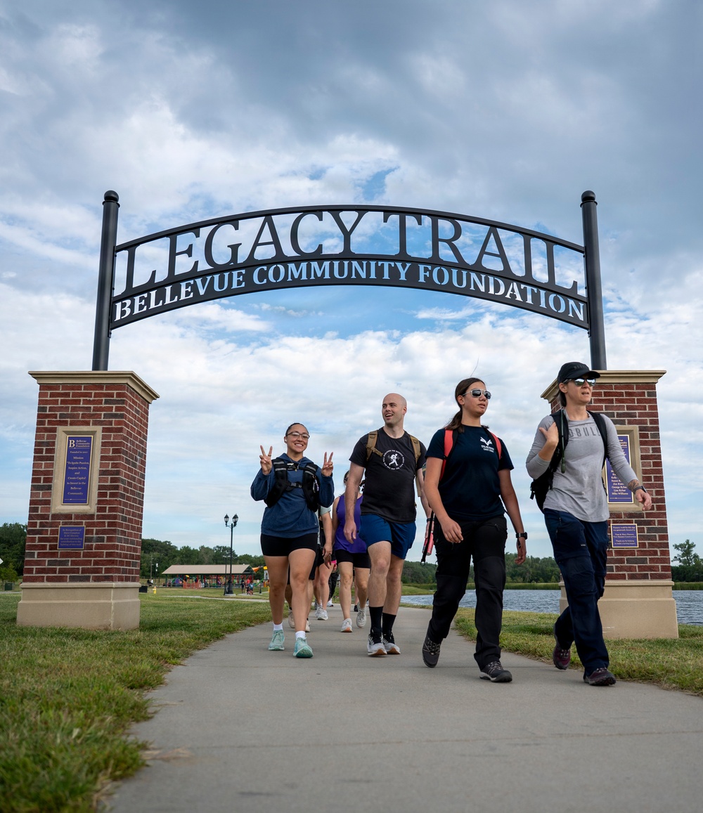 A group of people are walking along a paved trail beneath a decorative archway entrance gate reading 'Legacy Trail – Bellevue Community Foundation.' The walkers appear to be participating in an organized group walk or ruck march, as several are wearing backpacks. The setting is a well-maintained park with a lake, open green space, and a playground visible in the background.