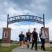 A group of people are walking along a paved trail beneath a decorative archway entrance gate reading 'Legacy Trail – Bellevue Community Foundation.' The walkers appear to be participating in an organized group walk or ruck march, as several are wearing backpacks. The setting is a well-maintained park with a lake, open green space, and a playground visible in the background.