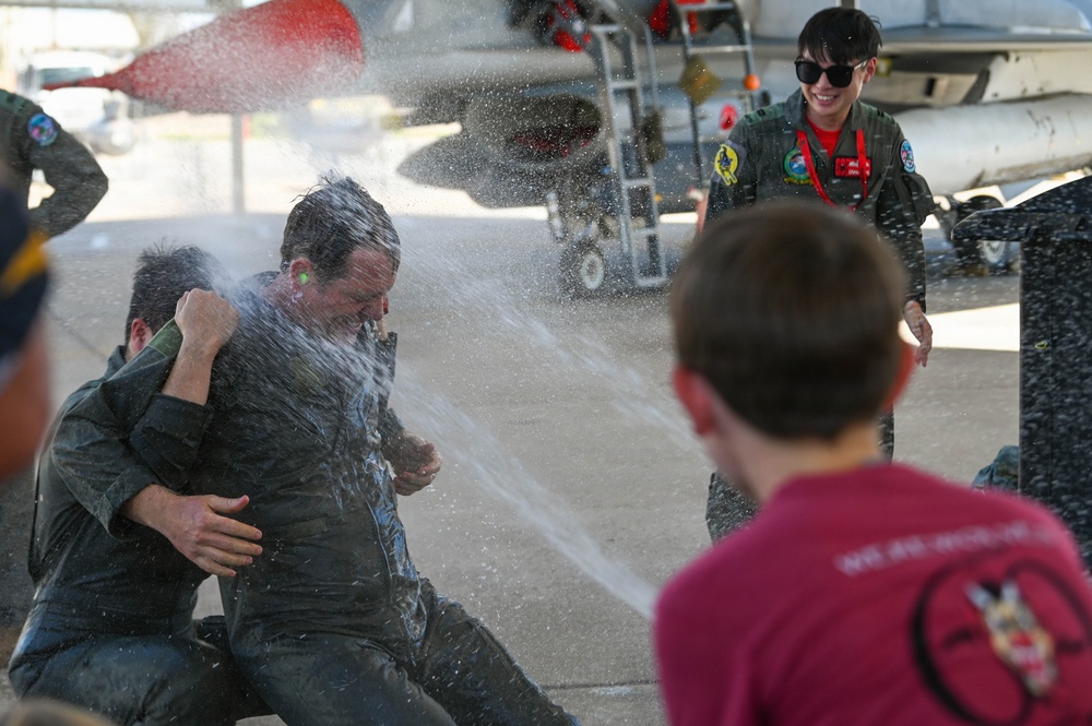 A military tradition known as a 'fini flight' or solo celebration is taking place on an airfield tarmac, where a person in a flight suit is being doused with water. A female pilot in a flight suit and sunglasses watches and smiles in the background, while a child in a red shirt participates in the water dousing. A military jet aircraft is visible in the background.
