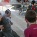 A military tradition known as a 'fini flight' or solo celebration is taking place on an airfield tarmac, where a person in a flight suit is being doused with water. A female pilot in a flight suit and sunglasses watches and smiles in the background, while a child in a red shirt participates in the water dousing. A military jet aircraft is visible in the background.