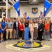 A group of approximately 25 civilians poses for a group photo inside an Air Force Materiel Command facility at Oklahoma City Air Logistics Complex (ALC). The group stands on a large floor seal of Air Force Materiel Command, surrounded by American flags and blue Air Force flags. The setting appears to be an industrial maintenance hangar or workshop floor.