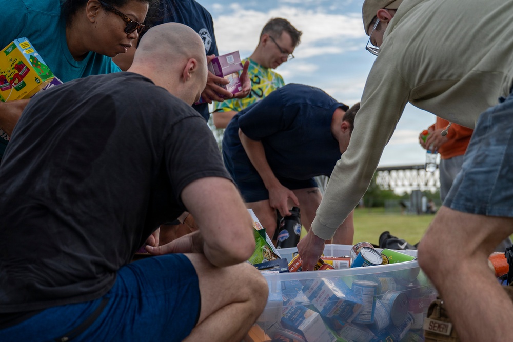 A group of people are gathered outdoors sorting through a large clear plastic bin filled with canned goods and packaged food items. The activity appears to be a food drive or community donation effort, with participants handling various boxed and canned products. The setting is an open grassy area with a bridge or industrial structure visible in the background.