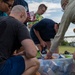 A group of people are gathered outdoors sorting through a large clear plastic bin filled with canned goods and packaged food items. The activity appears to be a food drive or community donation effort, with participants handling various boxed and canned products. The setting is an open grassy area with a bridge or industrial structure visible in the background.