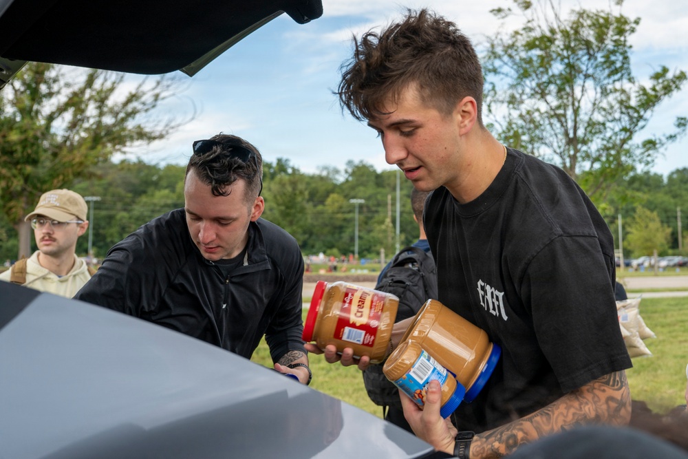 Two young men are handling large jars of peanut butter near the open trunk of a vehicle outdoors. A third person wearing a cap and glasses is visible in the background. The setting appears to be a park or open outdoor area with trees and green grass.