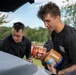 Two young men are handling large jars of peanut butter near the open trunk of a vehicle outdoors. A third person wearing a cap and glasses is visible in the background. The setting appears to be a park or open outdoor area with trees and green grass.