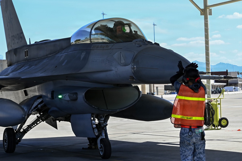 A military jet fighter, identifiable as an F-16 Fighting Falcon, is being marshaled on an airfield tarmac by a ground crew member wearing a high-visibility orange vest. A pilot is visible in the cockpit under the raised canopy. The scene appears to take place at a military air base, likely in a desert region based on the mountains and terrain visible in the background.