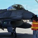 A military jet fighter, identifiable as an F-16 Fighting Falcon, is being marshaled on an airfield tarmac by a ground crew member wearing a high-visibility orange vest. A pilot is visible in the cockpit under the raised canopy. The scene appears to take place at a military air base, likely in a desert region based on the mountains and terrain visible in the background.