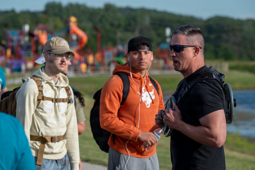 Three men are gathered outdoors near a body of water with a colorful playground structure visible in the background. The man on the right, wearing a black t-shirt, sunglasses, and carrying a weighted vest or pack, appears to be speaking or instructing the other two. The setting suggests an outdoor fitness or training event.