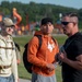 Three men are gathered outdoors near a body of water with a colorful playground structure visible in the background. The man on the right, wearing a black t-shirt, sunglasses, and carrying a weighted vest or pack, appears to be speaking or instructing the other two. The setting suggests an outdoor fitness or training event.