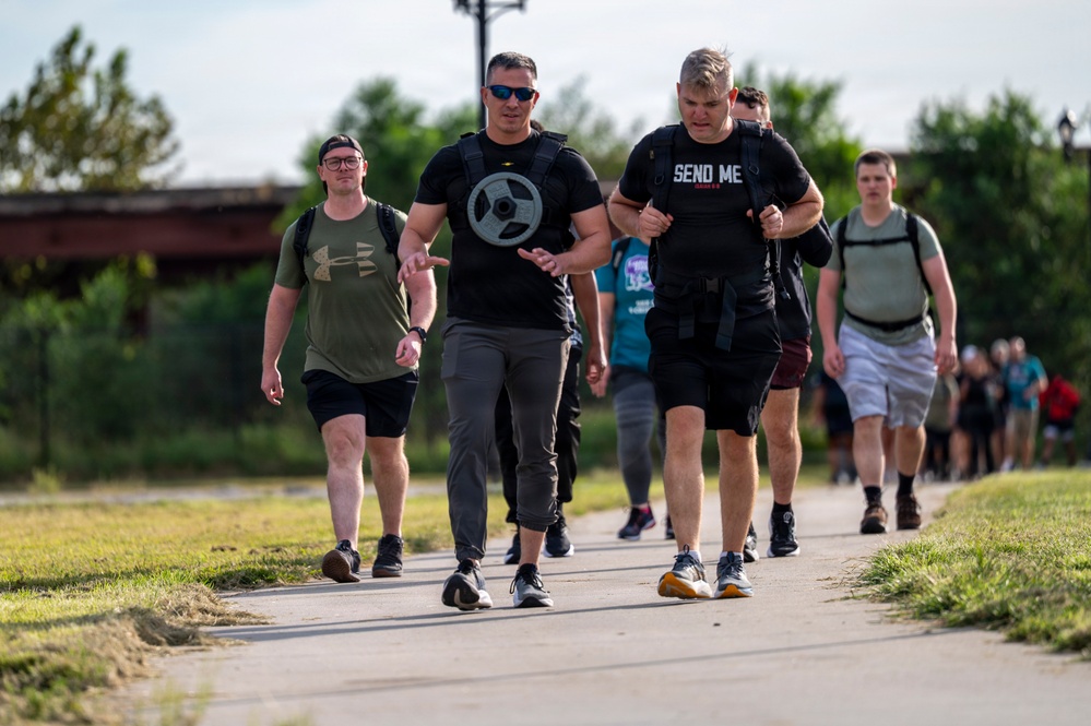 A group of people are participating in a rucking event or charity walk along a paved path in an outdoor setting. The lead participants are carrying weighted backpacks and a weight plate, suggesting a fitness or memorial challenge event. A large group of additional participants can be seen stretching into the background.