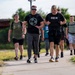 A group of people are participating in a rucking event or charity walk along a paved path in an outdoor setting. The lead participants are carrying weighted backpacks and a weight plate, suggesting a fitness or memorial challenge event. A large group of additional participants can be seen stretching into the background.