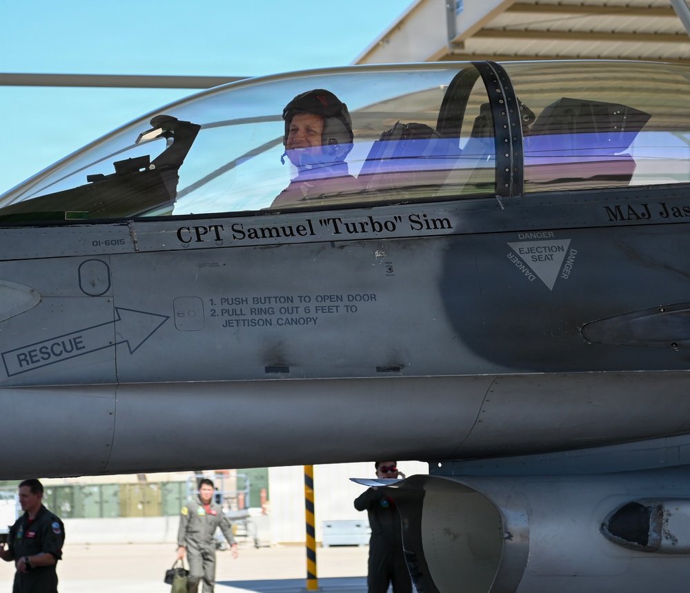 A military pilot is seated in the cockpit of an F-16 Fighting Falcon fighter jet on a flight line or tarmac. The aircraft bears the name 'CPT Samuel "Turbo" Sim' on the fuselage below the canopy, along with standard rescue and ejection seat markings. Ground crew personnel are visible in the background near a hangar.
