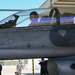 A military pilot is seated in the cockpit of an F-16 Fighting Falcon fighter jet on a flight line or tarmac. The aircraft bears the name 'CPT Samuel "Turbo" Sim' on the fuselage below the canopy, along with standard rescue and ejection seat markings. Ground crew personnel are visible in the background near a hangar.