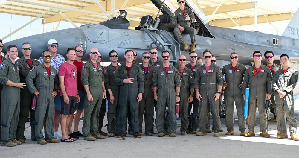 A large group of military personnel, predominantly wearing olive green flight suits, pose for a group photo in front of a gray fighter jet, likely an F-16, inside or near a hangar. One individual is seated on top of the aircraft's cockpit area. The setting appears to be a military air base, possibly during a special event or ceremony.