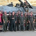 A large group of military personnel, predominantly wearing olive green flight suits, pose for a group photo in front of a gray fighter jet, likely an F-16, inside or near a hangar. One individual is seated on top of the aircraft's cockpit area. The setting appears to be a military air base, possibly during a special event or ceremony.