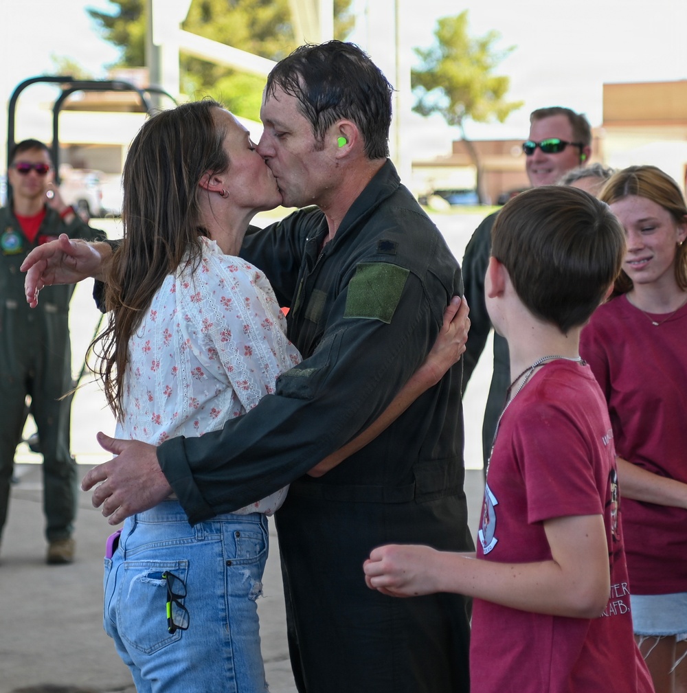A military pilot in a dark flight suit is being greeted with a kiss by a woman in a floral blouse and jeans, likely a homecoming or return from deployment ceremony. The pilot appears wet, possibly from a traditional post-flight dousing. Children and other personnel in flight suits are visible in the background.