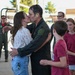 A military pilot in a dark flight suit is being greeted with a kiss by a woman in a floral blouse and jeans, likely a homecoming or return from deployment ceremony. The pilot appears wet, possibly from a traditional post-flight dousing. Children and other personnel in flight suits are visible in the background.