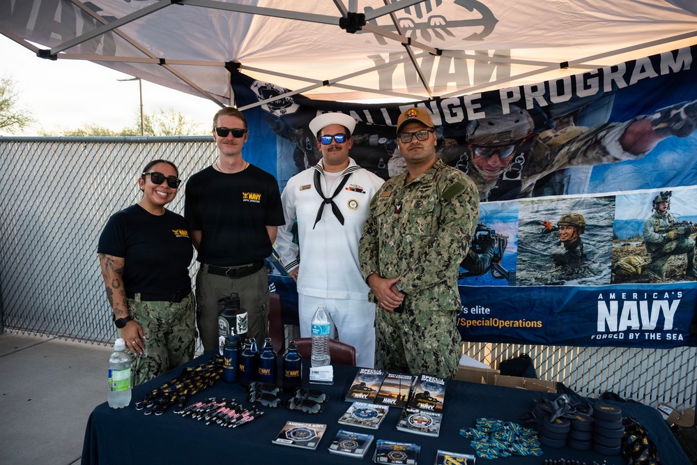 Four U.S. Navy personnel are posing at a recruitment/outreach booth under a canopy tent. The booth displays Navy branding, including an 'America's Navy - Forged by the Sea' banner and a 'Challenge Program' backdrop. A table in front is covered with promotional materials, branded merchandise, and informational brochures.
