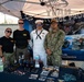 Four U.S. Navy personnel are posing at a recruitment/outreach booth under a canopy tent. The booth displays Navy branding, including an 'America's Navy - Forged by the Sea' banner and a 'Challenge Program' backdrop. A table in front is covered with promotional materials, branded merchandise, and informational brochures.